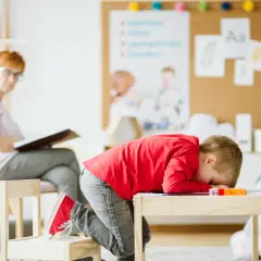 Chłopiec w czerwonej bluzie skulony nad stolikiem, z głową na rękach. Terapia dzieci zmagających się z chorobami psychicznymi.