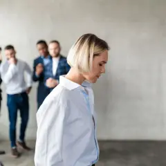 Kobieta odwrócona tyłem, podczas gdy trzej mężczyźni za jej plecami szepczą i wskazują na nią. Przykłady mobbingu w pracy.