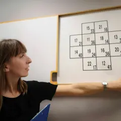 Kobieta wskazuje na tablicę z zadaniami, które mogą być częścią psychotestów. Pytania i odpowiedzi w formie liczb.