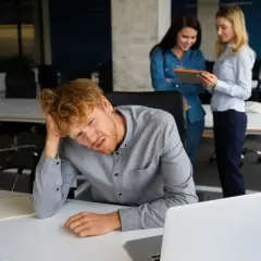Mężczyzna z rudymi włosami, lat 30, z ręką na głowie, wygląda na przygnębionego. Dwie kobiety rozmawiają w tle.