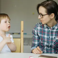 Chłopiec z otwartymi ustami i uniesionymi dłońmi, jakby walczył z utratą zdolności mówienia, patrzy na kobietę w okularach.