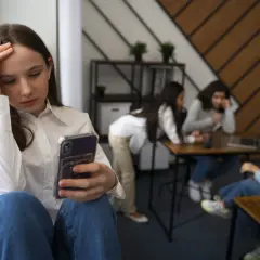 Dziewczyna z telefonem w dłoni, zmartwiona, odizolowana od grupy rówieśników. To obraz nieleczonej fobii społecznej.