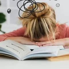 Dziewczynka z blond włosami w koku, zmęczona nauką, opiera głowę na otwartych książkach. Wokół jej głowy rysunki symbolizujące chaos i zmęczenie.