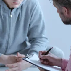 Mężczyzna w szarej bluzie rozmawia z terapeutą, który notuje na kartce. Rozmowa dotyczy nerwicy lękowej.