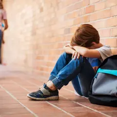 Chłopiec siedzi przy ceglanej ścianie, ukrywając twarz w dłoniach. W tle oddalają się inne dzieci. To obraz bullying po polsku.