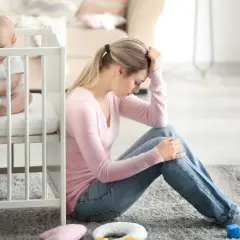 Zmęczona mama siedzi na podłodze obok łóżeczka, czując baby blues. Dziecko bawi się zabawką.