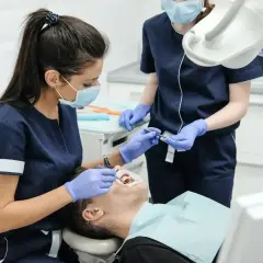 Dwie dentystki w maseczkach i rękawiczkach badają pacjenta w gabinecie stomatologicznym.