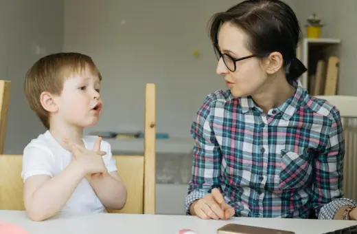 Chłopiec z otwartymi ustami i uniesionymi dłońmi, jakby walczył z utratą zdolności mówienia, patrzy na kobietę w okularach.