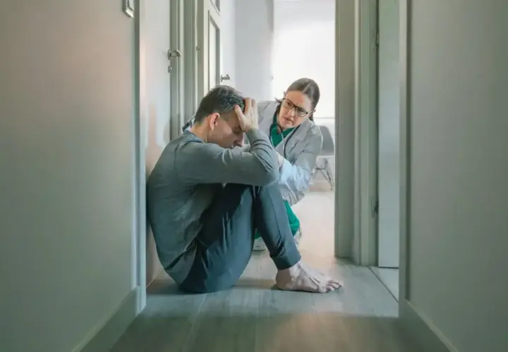 Mężczyzna w pozycji embrionalnej, z rękami na głowie, doświadcza objawów katatonii. Lekarka w białym fartuchu pochyla się nad nim, oferując wsparcie.