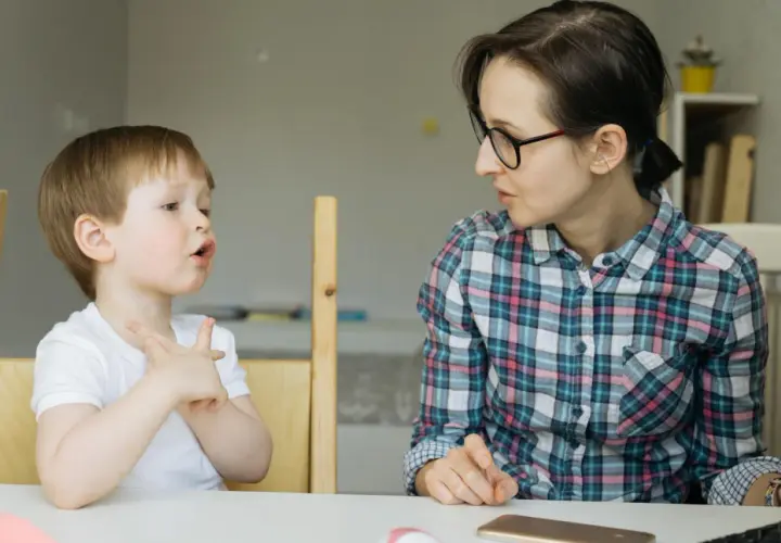 Chłopiec z otwartymi ustami i uniesionymi dłońmi, jakby walczył z utratą zdolności mówienia, patrzy na kobietę w okularach.