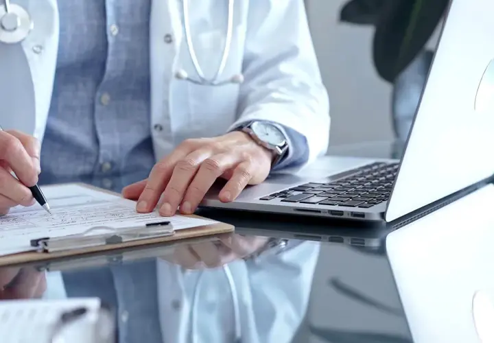 Lekarz w białym kitlu pisze na dokumentach, obok laptop. Szybka konsultacja psychiatryczna online.