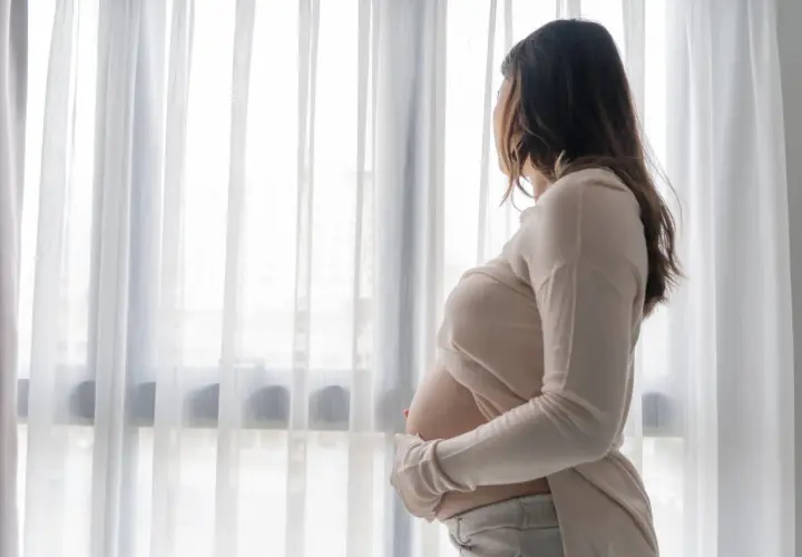 Kobieta w ciąży stoi przy oknie, patrząc na zewnątrz. Jej dłonie delikatnie obejmują brzuch, a w jej postawie można wyczuć smutek, sugerujący depresję ciążową.