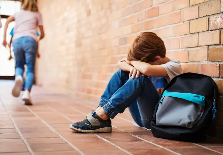 Chłopiec siedzi przy ceglanej ścianie, ukrywając twarz w dłoniach. W tle oddalają się inne dzieci. To obraz bullying po polsku.