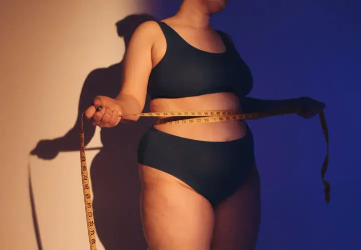 Osoba mierzy talię centymetrem. Ciemne światło podkreśla kształty, wywołując dyskomfort związany z body shaming.