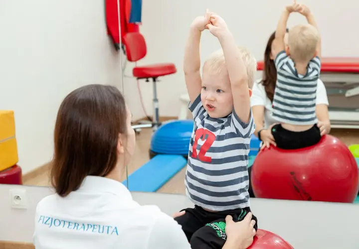 Fizjoterapeutka wspiera chłopca z zaburzeniami rozwojowymi podczas ćwiczeń na piłce rehabilitacyjnej.