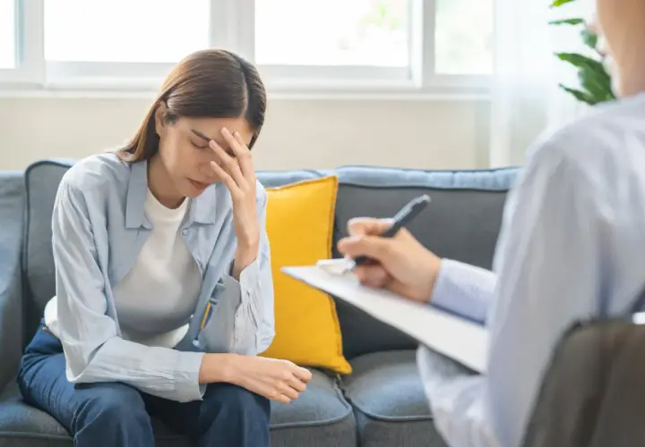 Kobieta z ręką na czole siedzi na kanapie, rozmawiając z terapeutą. Może to być rozmowa o diagnozie Aspergera.