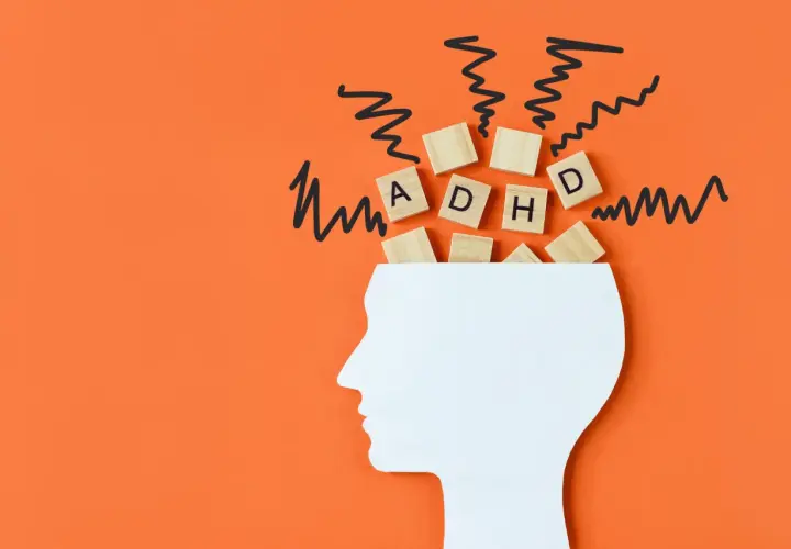 Biały profil głowy z literami "ADHD" na drewnianych kostkach i czarnymi, zygzakowatymi liniami symbolizującymi chaos. Czy ADHD można nabyć?