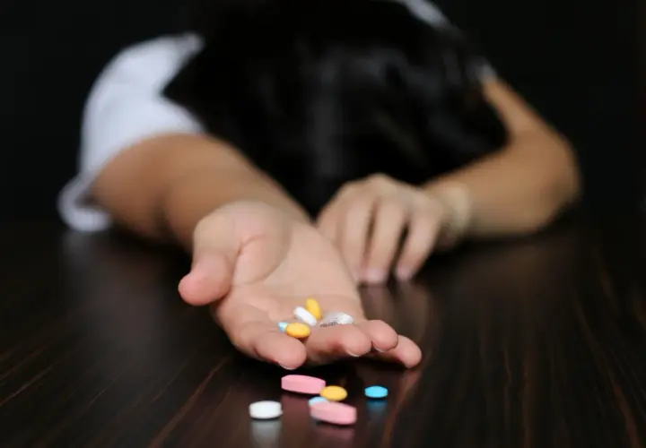 Otwarta dłoń z garścią tabletek, w tym Xanax, na co wskazują różne kolory i kształty. Osoba leży bezwładnie na stole.