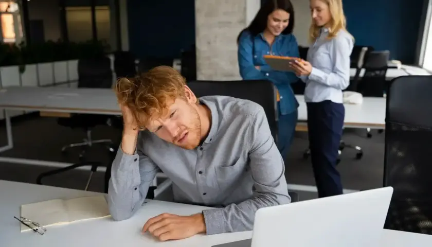 Mężczyzna z rudymi włosami, lat 30, z ręką na głowie, wygląda na przygnębionego. Dwie kobiety rozmawiają w tle.