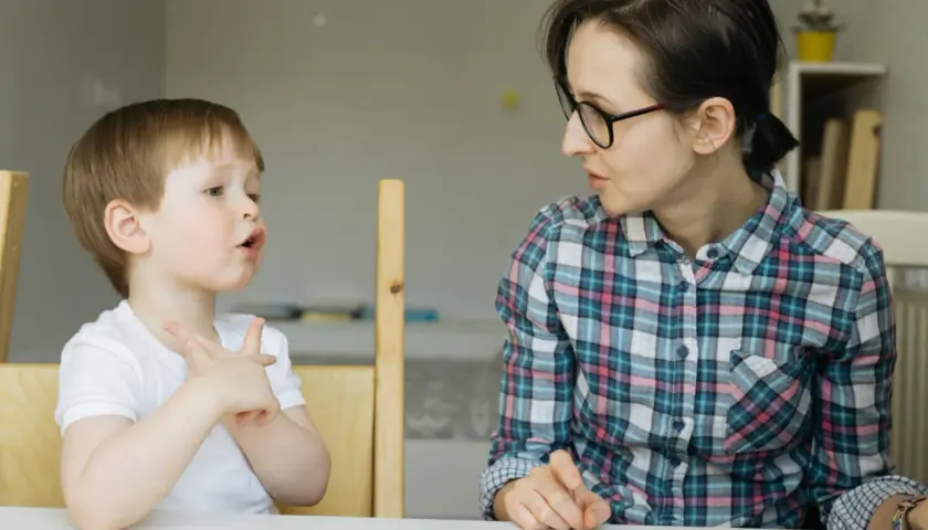 Chłopiec z otwartymi ustami i uniesionymi dłońmi, jakby walczył z utratą zdolności mówienia, patrzy na kobietę w okularach.