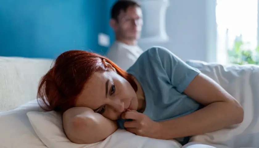 Smutna kobieta leży w łóżku, zamyślona. W tle niewyraźny mężczyzna. Obraz symbolizuje trudne relacje i toksyczny partner cechy.