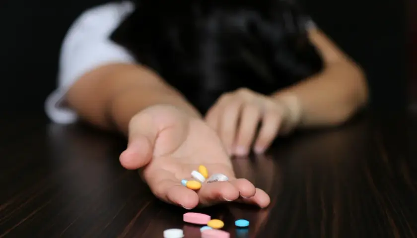 Otwarta dłoń z garścią tabletek, w tym Xanax, na co wskazują różne kolory i kształty. Osoba leży bezwładnie na stole.