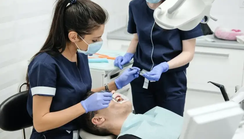 Dwie dentystki w maseczkach i rękawiczkach badają pacjenta w gabinecie stomatologicznym.