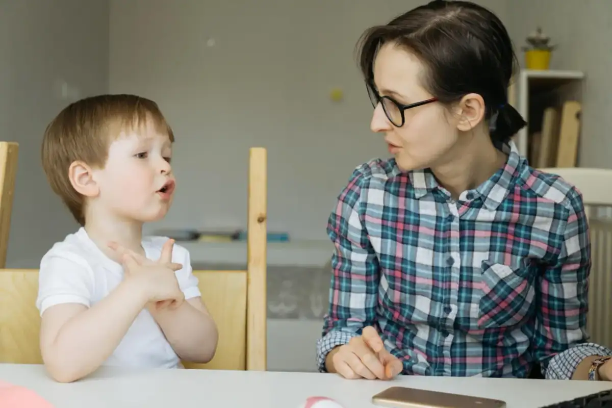 Chłopiec z otwartymi ustami i uniesionymi dłońmi, jakby walczył z utratą zdolności mówienia, patrzy na kobietę w okularach.