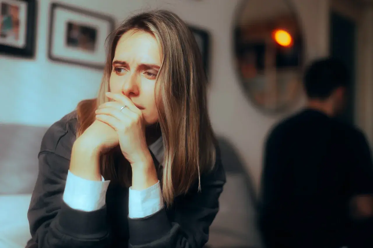 Smutna kobieta zamyślona, z rękami przy twarzy. W tle niewyraźna postać mężczyzny. Obraz symbolizuje obojętność w związku.