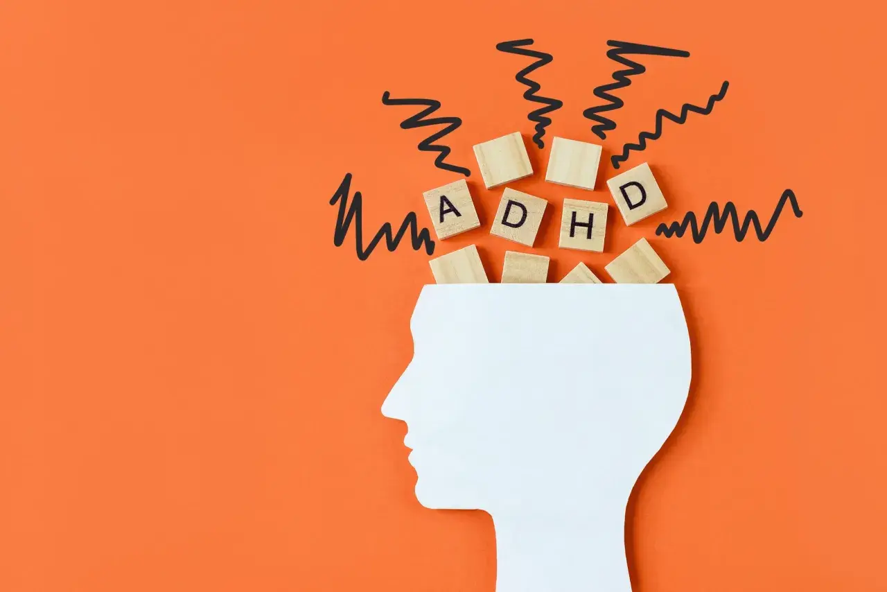 Biały profil głowy z literami "ADHD" na drewnianych kostkach i czarnymi, zygzakowatymi liniami symbolizującymi chaos. Czy ADHD można nabyć?