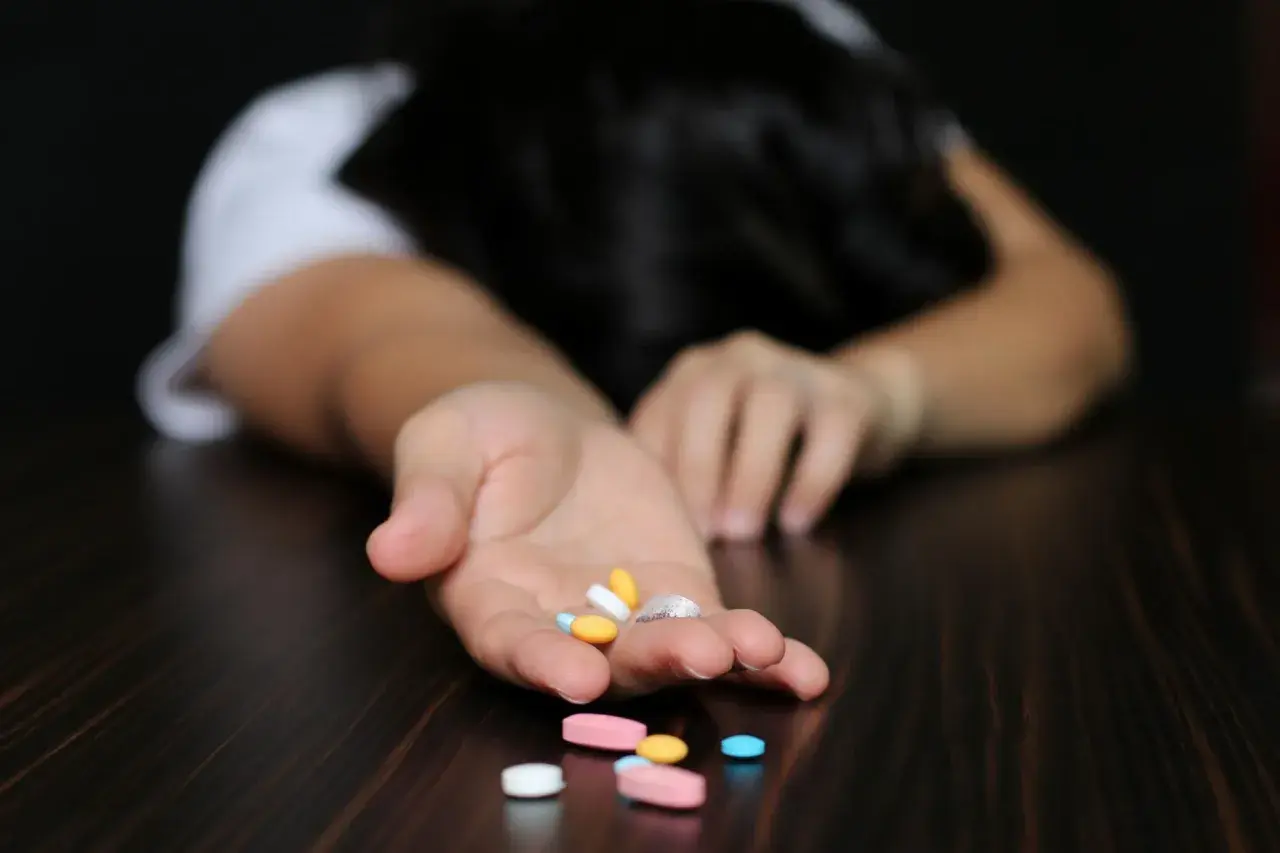 Otwarta dłoń z garścią tabletek, w tym Xanax, na co wskazują różne kolory i kształty. Osoba leży bezwładnie na stole.